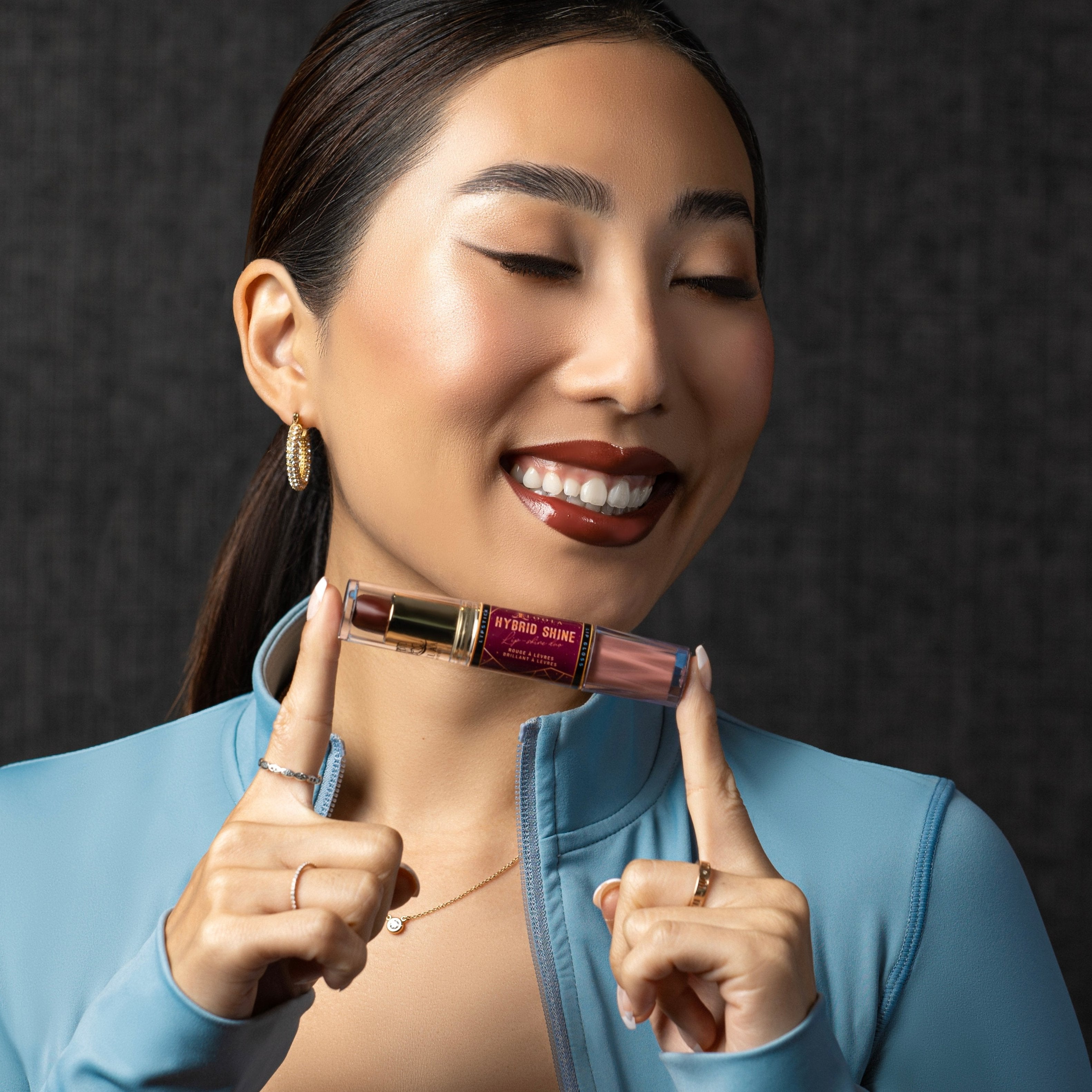 Model smilling and holdine the Cannes Hybrid Shine - Lipgloss and Lipstick Duo - its a brown lipstick and a gloss